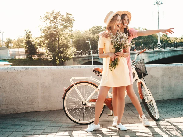 Two young beautiful smiling hipster woman in trendy summer sundress. Sexy carefree women posing on the street background in hats. Positive models at embankment at sunset near bicycle