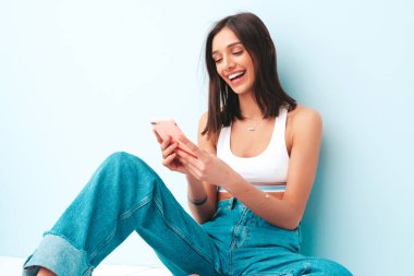 Beyaz formalı bluz ve kot pantolon giymiş güzel bir kadın. Stüdyoda açık mavi duvarın yanında poz veren neşeli, seksi bir model. Cep telefonu ekranına bakıyor.