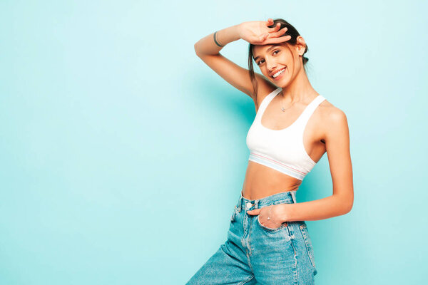 Beautiful smiling woman dressed in white jersey top shirt and jeans. Sexy carefree cheerful model enjoying her morning. Adorable and positive female posing near blue wall in studio
