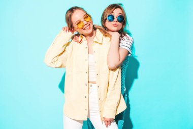 Moda kıyafetleri içinde gülümseyen iki güzel sarışın hippi kadın. Stüdyoda mavi duvarın yanında poz veren kaygısız seksi kadınlar. Moda ve pozitif modeller eğleniyor.