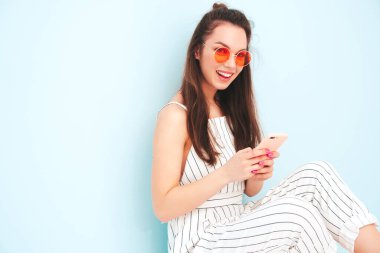 Son moda yaz hippileri içinde gülümseyen genç bir bayan tulum giyiyor. Stüdyoda açık mavi duvarın yanında poz veren kaygısız seksi kadın. Pozitif model içeride eğleniyor. Akıllı telefon uygulamaları kullanmaktan keyif alıyorum.