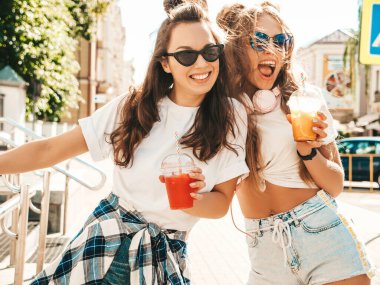 Moda kıyafetleri içinde gülümseyen iki güzel hippi kadın. Dışarıda poz veren kaygısız kadınlar. Pozitif modeller, plastik bardakta pipetle taze kokteyl içiyorlar ve içiyorlar.