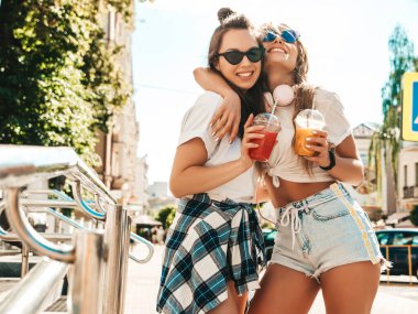 Moda kıyafetleri içinde gülümseyen iki güzel hippi kadın. Dışarıda poz veren kaygısız kadınlar. Pozitif modeller, plastik bardakta pipetle taze kokteyl içiyorlar ve içiyorlar.