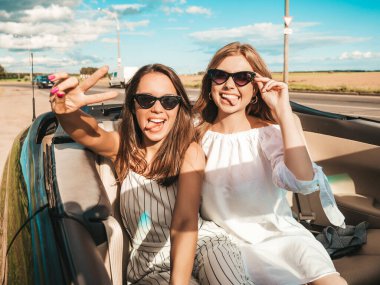 Üstü açık arabadaki iki güzel ve gülümseyen hippi kadının portresi. Cabriolet kullanan kaygısız seksi kadınlar. Pozitif modeller dışarıda güneş gözlüğü takıp eğleniyorlar. Yaz günlerinin tadını çıkarıyorlar.