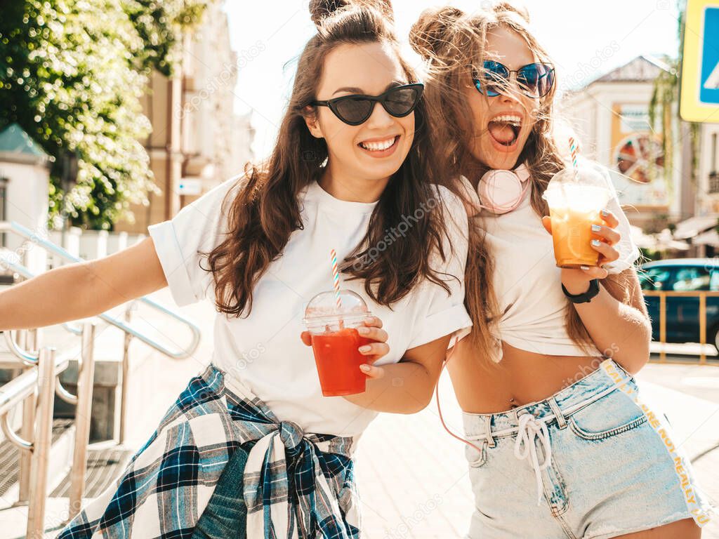 Two young beautiful smiling hipster female in trendy summer clothes. Carefree women posing outdoors.Positive models holding and drinking fresh cocktail smoothie drink in plastic cup with straw