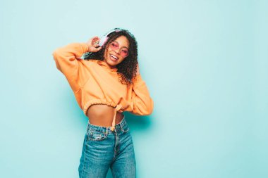 Afro kıvırcık saçlı güzel bir zenci kadın. Turuncu kapüşonlu ve kot pantolonlu bir model. Kablosuz kulaklıkla müzik dinleyen kaygısız bir kadın.