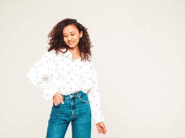 Saçları kıvırcık, güzel siyah bir kadın. Beyaz gömlekli, kotlu, gülen bir model. Stüdyoda gri arka planda poz veren kaygısız seksi kadın. Bronzlaşmış ve neşeli 