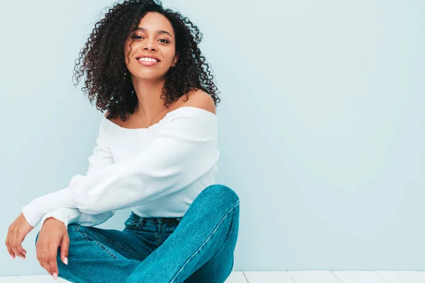 Beautiful black woman with afro curls hairstyle.Smiling model in sweater and trendy jeans clothes. Sexy carefree female sitting near light blue wall in studio. Tanned and cheerful