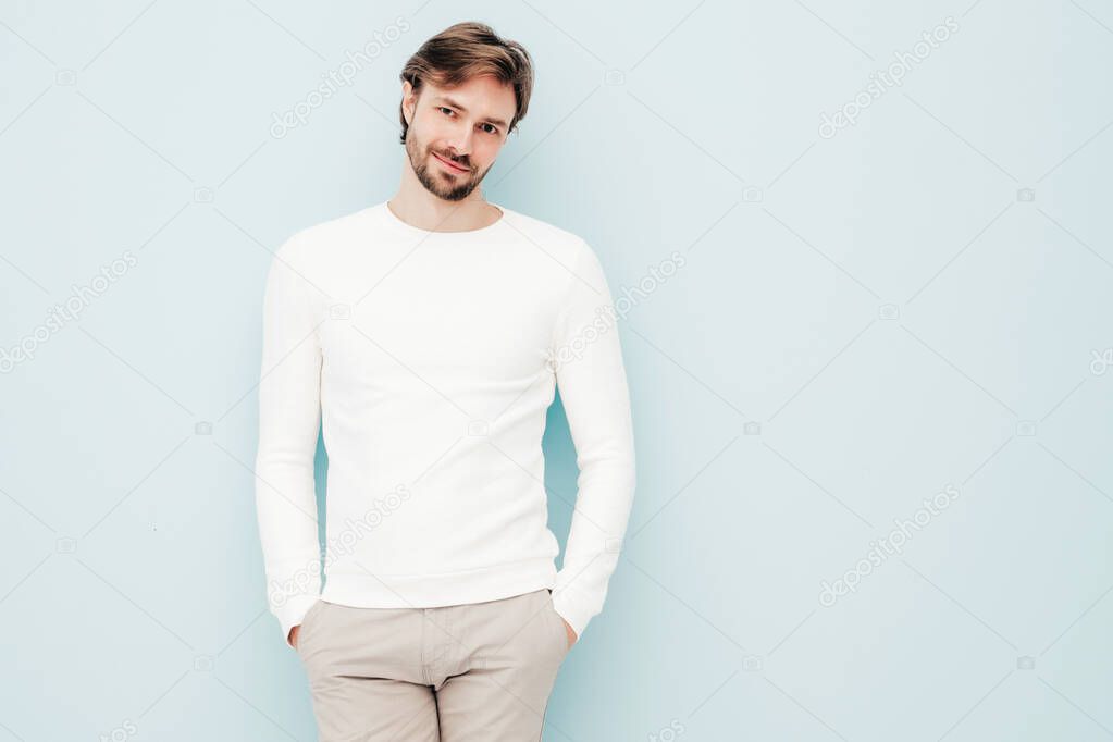 Portrait of handsome smiling hipster lumbersexual businessman model wearing casual white sweater and trousers. Fashion stylish man sitting against light blue wall in studio