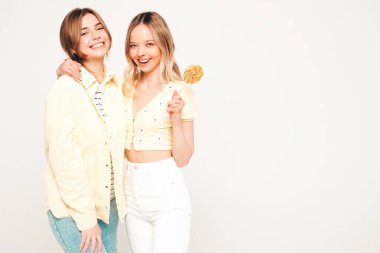 Moda kıyafetleri içinde gülümseyen iki güzel sarışın hippi kadın. Stüdyoda poz veren kaygısız seksi kadınlar. Moda ve pozitif modeller şekerleme lolipoplarıyla eğleniyorlar