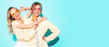 Moda kıyafetleri içinde gülümseyen iki güzel sarışın hippi kadın. Stüdyoda mavi duvarın yanında poz veren kaygısız seksi kadınlar. Moda ve pozitif modeller şekerleme lolipoplarıyla eğleniyorlar