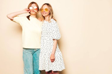 Moda kıyafetleri içinde gülümseyen iki güzel sarışın hippi kadın. Stüdyoda duvarın yanında poz veren kaygısız seksi kadınlar. Moda ve pozitif modeller güneş gözlükleriyle eğleniyor.