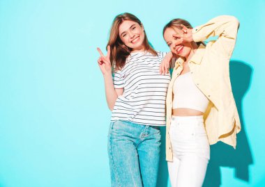 Moda kıyafetleri içinde gülümseyen iki güzel sarışın hippi kadın. Stüdyoda mavi duvarın yanında poz veren kaygısız seksi kadınlar. Moda ve pozitif modeller eğleniyor.