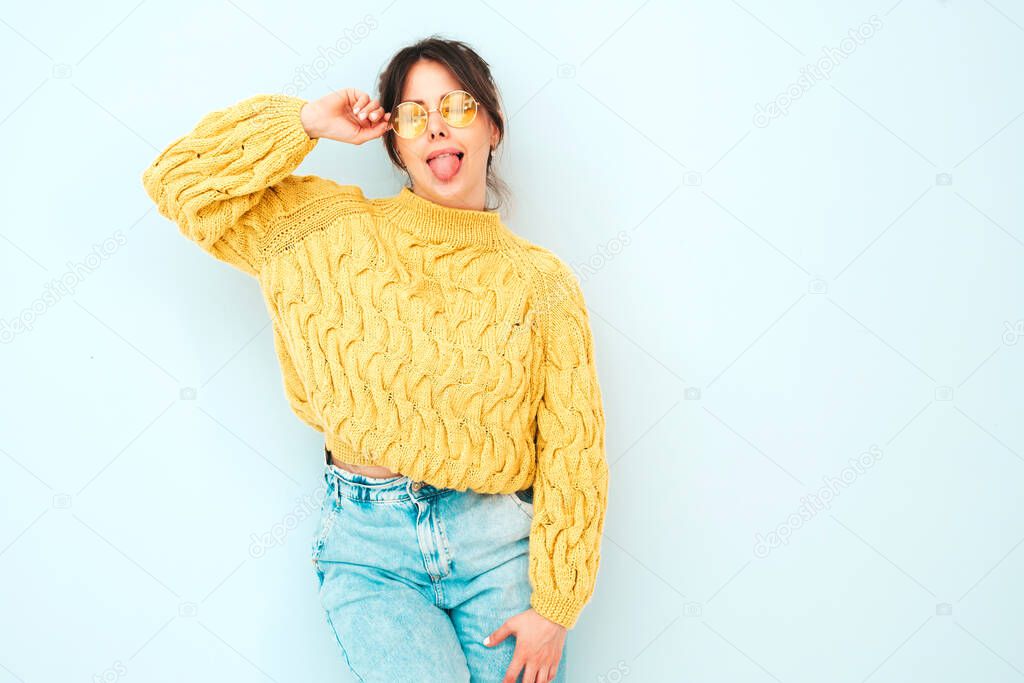 Young beautiful smiling female in trendy summer yellow hipster sweater and  jeans. Sexy carefree woman posing near light blue wall in studio. Positive model having fun indoors. Shows tongue