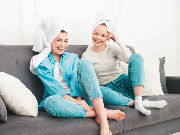 Two young beautiful smiling women sitting at the sofa. Sexy carefree models posing indoors in posh apartment or hotel room. They doing beauty treatments at home in towels on heads