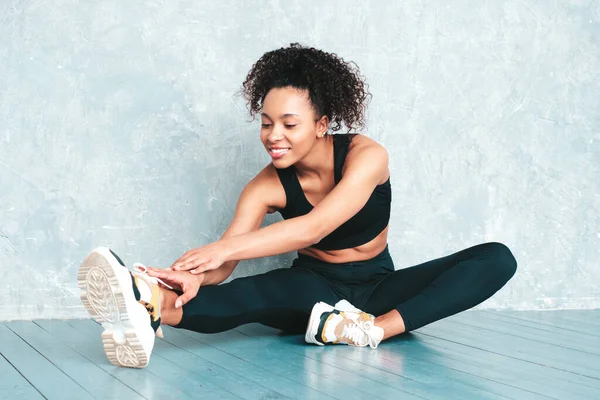 Portrait of fitness smiling black woman in sports clothing with afro curls hairstyle.She wearing sportswear.Young beautiful model stretching out before training.Female sitting in studio near gray wall