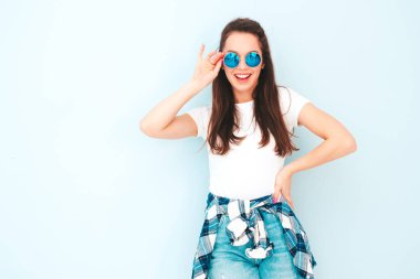 Yeni moda hippi kıyafetleri içinde gülümseyen güzel bir kadın. Stüdyoda açık mavi duvarın yanında poz veren kaygısız seksi kadın. Pozitif model içeride eğleniyor. Neşeli ve mutlu güneş gözlükleriyle