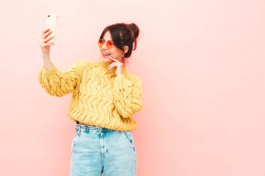 Son moda sarı hippi süveter ve kot pantolonlu güzel, gülümseyen bir bayan. Stüdyoda pembe duvarın yanında poz veren kaygısız seksi kadın. Pozitif model eğleniyor. Selfie çekiyor.