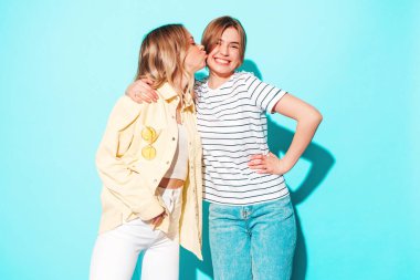 Moda kıyafetleri içinde gülümseyen iki güzel sarışın hippi kadın. Stüdyoda mavi duvarın yanında poz veren kaygısız seksi kadınlar. Moda ve pozitif modeller eğleniyor. Arkadaşını yanağından öpüyor. 