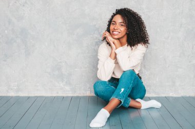 Saçları kıvırcık, güzel siyah bir kadın. Moda kotlar ve güneş gözlükleriyle gülümseyen bir model. Stüdyoda gri duvarın yanında oturan kaygısız seksi kadın.