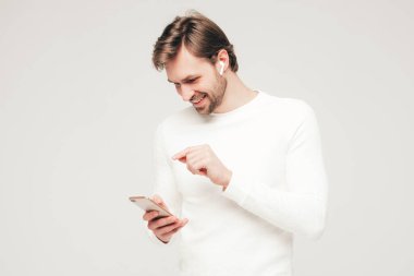 Handsome smiling hipster lumbersexual businessman model wearing white casual sweater and trousers.Fashion stylish man posing against gray background in studio. Holding smartphone in hands.In earphones clipart