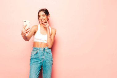 Beyaz formalı bluz ve kot pantolon giymiş güzel bir kadın. Seksi kaygısız neşeli model içeride eğleniyor. Stüdyoda pembe duvarın yanında poz veren sevimli ve pozitif kadın. Selfie çekiyorum.