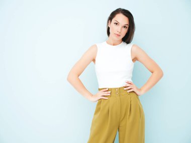 Moda kıyafetleri içinde gülümseyen genç ve güzel bir bayan. Stüdyoda açık mavi duvarın yanında poz veren kaygısız seksi kadın. Pozitif model içeride eğleniyor. Neşeli ve mutlu