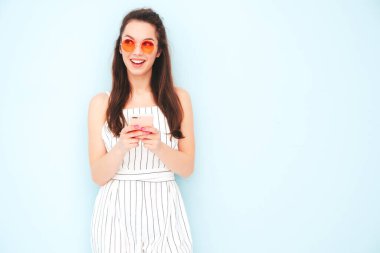 Son moda yaz hippileri içinde gülümseyen genç bir bayan tulum giyiyor. Stüdyoda açık mavi duvarın yanında poz veren kaygısız seksi kadın. Pozitif model içeride eğleniyor. Akıllı telefon uygulamaları kullanmaktan keyif alıyorum.