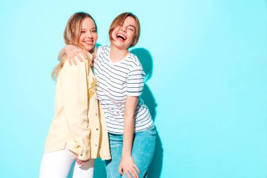 Moda kıyafetleri içinde gülümseyen iki güzel sarışın hippi kadın. Stüdyoda mavi duvarın yanında poz veren kaygısız seksi kadınlar. Moda ve pozitif modeller eğleniyor. 