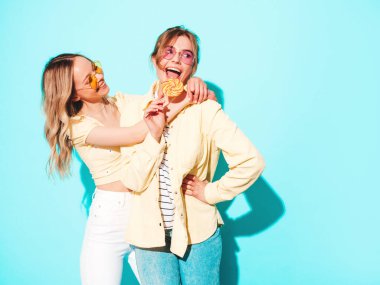 Moda kıyafetleri içinde gülümseyen iki güzel sarışın hippi kadın. Stüdyoda mavi duvarın yanında poz veren kaygısız seksi kadınlar. Moda ve pozitif modeller eğleniyor. 