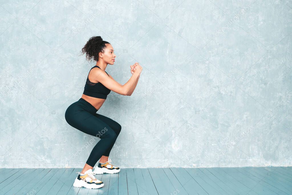 Fitness smiling black woman in sports clothing with afro curls hairstyle.She doing squats. Young beautiful model with perfect tanned body.Female in studio near gray wall