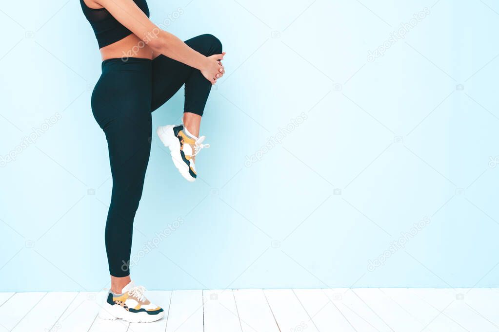 Fitness smiling black woman in sports clothing with afro curls hairstyle.She wearing sportswear.Young model stretching out legs before training.Female sitting in studio near blue wall. No face