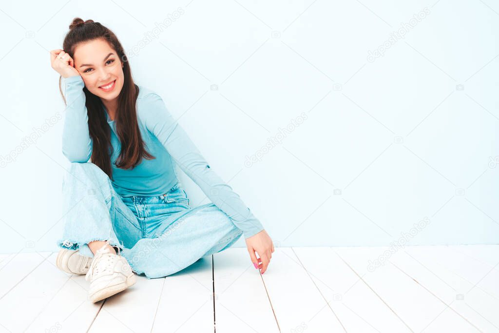 Young beautiful smiling female in trendy summer hipster clothes. Sexy carefree woman sitting near light blue wall in studio. Positive model having fun indoors. Cheerful and happy 