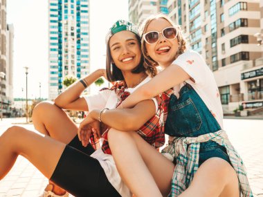 Renkli kaykaylı, gülümseyen iki genç bayan. Yazlık hippi elbiseli kadınlar arka planda oturuyor. Pozitif modeller eğleniyor ve deliriyor.