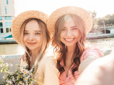 İki genç, güzel, gülümseyen yeniyetme kadın, modaya uygun bir yaz elbisesi içinde. Sokakta şapkalarla poz veren kaygısız seksi kadınlar. Sunset 'te pozitif modeller var. Selfii alıyorum. Çiçekleri tutuyorum.