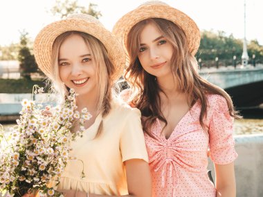 İki genç, güzel, gülümseyen yeniyetme kadın, modaya uygun bir yaz elbisesi içinde. Sokakta şapkalarla poz veren kaygısız seksi kadınlar. Sunset 'te olumlu modeller var. Çiçek tutuyorlar.