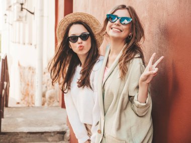 Son moda beyaz süveter ve palto giymiş iki güzel, gülümseyen hippi kadın. Sokakta şapkalarla poz veren kaygısız seksi kadınlar. Pozitif modeller dışarıda eğleniyor. Güneş gözlükleriyle sarılmak.