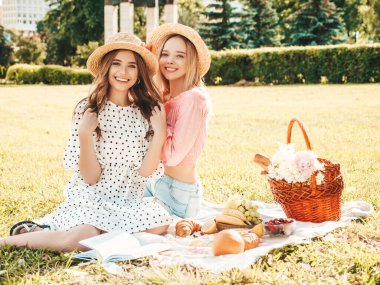Moda elbisesi ve şapkası içinde iki güzel bayan. Dışarıda piknik yapan kaygısız kadınlar. Çimlerin üzerinde oturmuş meyve ve peynir yiyen pozitif modeller.