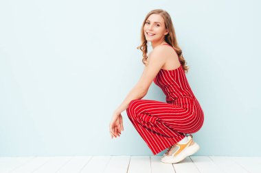 Yeni moda kırmızı takım elbiseli güzel, gülümseyen bir kadının portresi. Stüdyoda açık mavi duvarın yanında poz veren kaygısız seksi kadın. Pozitif model içeride eğleniyor. Neşeli ve mutlu