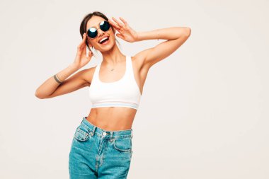Beautiful smiling woman dressed in white jersey top shirt and jeans. Sexy carefree cheerful model having fun indoors.Adorable and positive female posing on grey background in studio in sunglasses clipart