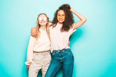 Moda kıyafetleri içinde iki güzel, gülümseyen uluslararası hippi kadın. Stüdyoda mavi duvarın yanında poz veren kaygısız seksi kadınlar. Pozitif modeller eğleniyor. Arkadaşlık kavramı