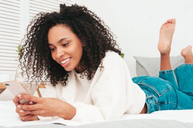 Saçları kıvırcık, güzel, siyah bir kadın. Seksi gülümseyen manken sabahları yatak odasındaki beyaz iç kısımda yatıyor. Akıllı telefon ekranına bakıyorum. Akıllı telefon uygulamaları kullanıyorum.