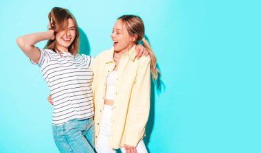 Moda kıyafetleri içinde gülümseyen iki güzel sarışın hippi kadın. Stüdyoda mavi duvarın yanında poz veren kaygısız seksi kadınlar. Moda ve pozitif modeller eğleniyor.