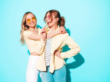 Moda kıyafetleri içinde gülümseyen iki güzel sarışın hippi kadın. Stüdyoda mavi duvarın yanında poz veren kaygısız seksi kadınlar. Moda ve pozitif modeller güneş gözlükleriyle eğleniyor.