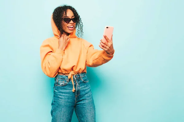 Beautiful black woman with afro curls hairstyle.Smiling model in orange hoodie and trendy jeans clothes. Sexy carefree female posing near blue wall in studio in sunglasses.Taking selfie