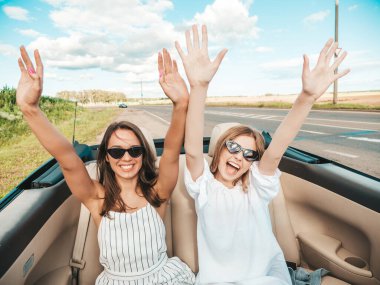 Üstü açık arabadaki iki güzel ve gülümseyen hippi kadının portresi. Cabriolet kullanan kaygısız seksi kadınlar. Pozitif modeller dışarıda güneş gözlüğü takıp eğleniyorlar. Yaz günlerinin tadını çıkarıyorlar.