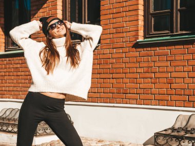 Güzel gülümseyen modelin portresi. Sıcak beyaz kazak ve bere giymiş bir kadın. Gün batımında sokakta poz veriyor.