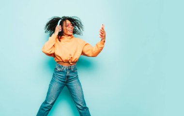 Afro kıvırcık saçlı güzel bir zenci kadın. Turuncu kapüşonlu ve kot pantolonlu bir model. Kablosuz kulaklıkla müzik dinleyen kaygısız bir kadın.