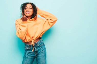 Afro kıvırcık saçlı, turuncu kapüşonlu ve kotlu güzel siyah kadın. Stüdyoda mavi duvarın yanında güneş gözlükleriyle poz veren seksi kadın. Hava öpücüğü veriyor.