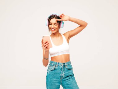 Kot pantolon giymiş, gülümseyen güzel bir kadın. Kablosuz kulaklıkla müzik dinleyen seksi tasasız model. Stüdyoda gri arka planda poz veren sevimli ve pozitif bir kadın. Akıllı telefon tutuluyor